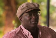 Journalist Thomas Dixon Calls for Genuine Reconciliation Beyond Political Settlement Man wearing a brown flat cap and patterned pink shirt sits at a wooden table outdoors, looking to the side with glasses on.