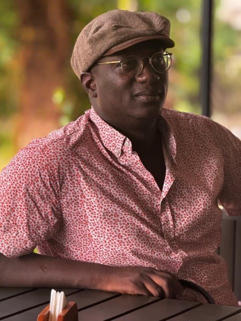 Guild of Newspapers Editors, Thomas Josephus Dixon Man wearing a brown flat cap and patterned pink shirt sits at a wooden table outdoors, looking to the side with glasses on.