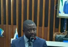 Attorney General Secures Parliamentary Approval for African Human Rights Court Protocol Man in a blue suit speaks at a wooden podium in a formal meeting room, with a microphone in front.