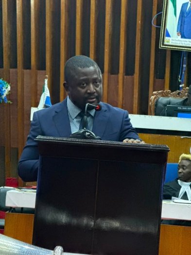 Attorney General and Minister of Justice, Alpha Sesay Esq. Man in a blue suit speaks at a wooden podium in a formal meeting room, with a microphone in front.
