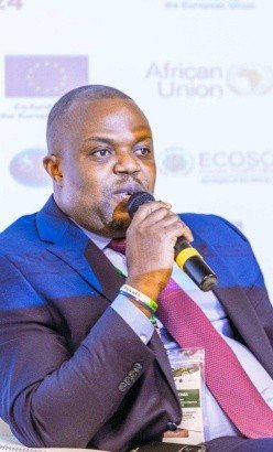 Chief Executive of Campaign for Human Rights and Development International (CHRDI), Abdul Fatoma Man in a blue suit and pink tie speaks into a microphone at a conference, wearing a lanyard; blurred backdrop shows EU and African Union logos.