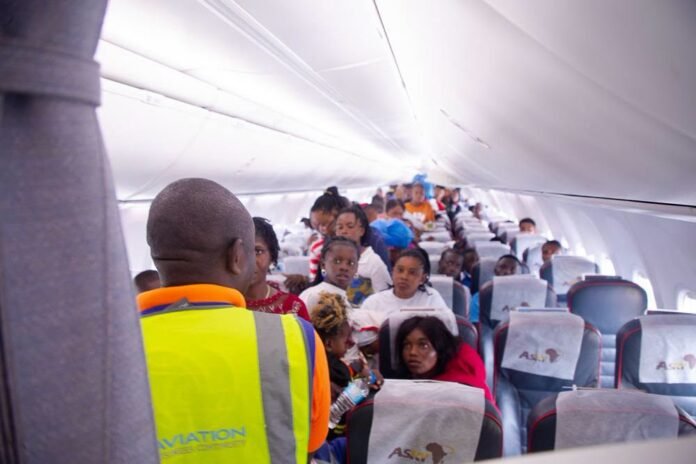 Flight attendant in a neon safety vest standing at the front of a crowded airplane cabin, passengers seated and looking ahead