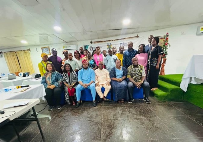 Group of diverse adults posing for a group photo in an indoor meeting hall with tables and chairs nearby.