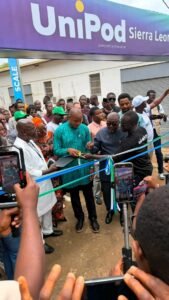 | The Calabash Newspaper A group of people at a ribbon-cutting ceremony under a large UniPod Sierra Leone banner, with a man cutting the blue ribbon while others watch and film.