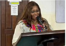 HRC-SL Submits Position Paper on Constitutional Amendment Bill to Parliament Woman speaking at a wooden podium with a microphone, wearing glasses and a beige blouse with a colorful beaded neckline.