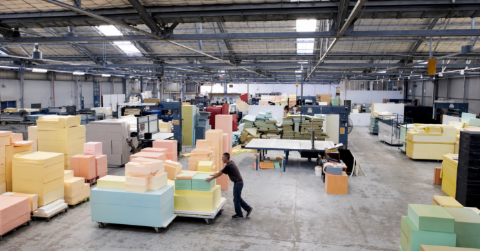 Factory floor with stacks of colorful foam blocks on pallets and a worker pushing a cart to the left side of the image.