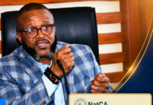 NatCA Reassures Public and Strengthens Response to Rising Cyber Threats A man wearing glasses and a blue plaid blazer speaks into a microphone at a briefing, with a NatCA public notice displayed beside him on the desk edge.