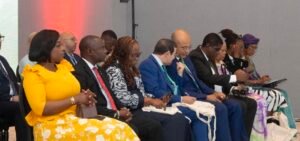 | The Calabash Newspaper People seated in a row at a formal event, wearing business attire; one woman in a bright yellow dress foreground.