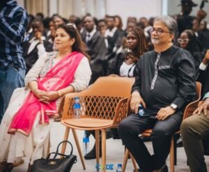 Audience seated at a formal event: woman in a pink saree on the left and a man in a black traditional outfit on the right, with others blurred in the background.