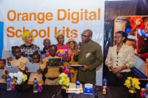 Group of children and adults standing behind a table with Orange Digital School kits on display; a man speaks into a microphone while others hold tablets, with a banner reading 'Orange Digital School kits' in the background.