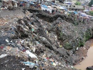 Waste-covered hillside dump next to crowded shacks, with a muddy water channel at the bottom.