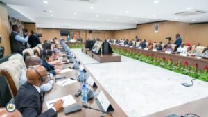 A formal conference with officials seated around a long U-shaped table, microphones, water bottles, and a central podium.