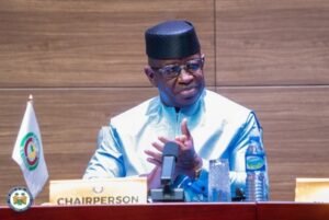 Chairperson in blue traditional attire speaks at a desk with a microphone, CHAIRPERSON sign, and a flag nearby.