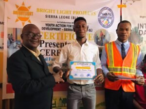 Man in a white shirt receives a certificate from a suited man, with a man in an orange safety vest on the right at a ceremony in front of a banner.
