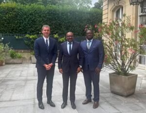 Three men in dark suits posing together outdoors in a courtyard with potted plants and a hedge in the background.