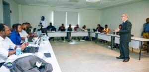Diverse professionals seated around U-shaped tables in a conference room, listening to a male presenter in a suit at the front.