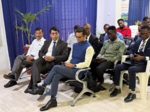 Group of professionally dressed men seated in a conference room with blue curtains, listening to a meeting; one man in a blue vest and yellow shirt sits in front, others in suits nearby; a potted plant is visible in the background.