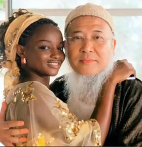 Young Black woman in a gold-embellished sheer outfit hugs an older East Asian man with a white beard and glasses.