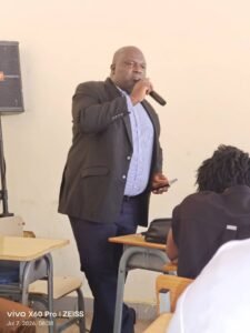 Lead Investigator for the Pepsin study, Dr Edries Tejan, presenting the findings | The Calabash Newspaper Man in a dark blazer speaks into a handheld microphone at the front of a classroom, addressing students seated at desks.