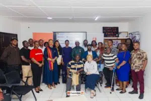 Group photo of diverse participants at a foundation event in a bright meeting room, with two people seated at the front and a Foundation banner in the background.