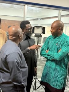 | The Calabash Newspaper Three men in a modern indoor workspace are in discussion: one man in a black shirt points as the others listen.