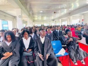 | The Calabash Newspaper Graduates in black gowns seated in a bright hall during a graduation ceremony, some checking phones, with a red carpet on the floor.