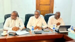 Three men in white shirts sit at a conference table with documents, laptops, and water bottles during a meeting.