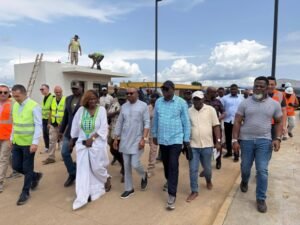| The Calabash Newspaper Group of officials walking together on a construction site, led by a woman in a white traditional dress amid workers and equipment in background.