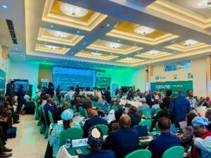 Large conference hall filled with attendees seated at round tables facing a stage with screens and banners.