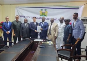 | The Calabash Newspaper Group of officials in a conference room pose as two men exchange a folder in front of a Ministry banner.