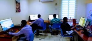Five people seated at computer workstations in a classroom computer lab working on desktop PCs.