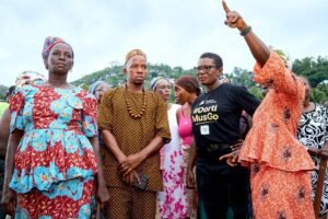 Group of diverse adults gathered outdoors, with one woman in a black '#DortiMusGo' shirt raising her arm and speaking to the crowd.
