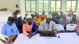 Group of men in colorful traditional attire seated at a long table in a bright room during a community meeting; one man uses a tablet at the center of the table.