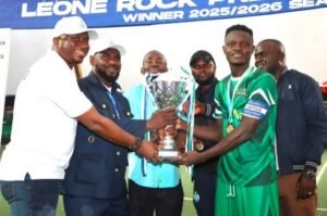| The Calabash Newspaper Group of players and officials pass a large trophy on a stadium field after a win, smiling for the camera.
