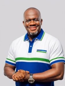 | The Calabash Newspaper Smiling man in a white polo with blue and green stripes, standing against a light gray background with hands clasped.
