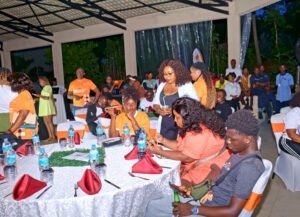 People seated at decorated banquet tables during an outdoor event, many wearing orange shirts, with bottled water and red napkins on the tables.