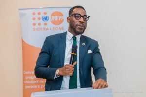 Man in a teal suit and green tie speaks into a microphone at a podium, with a UNFPA Sierra Leone banner in the background.