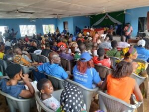 Community gathering in a brightly colored hall with people seated in plastic chairs facing a stage where speakers address the crowd.