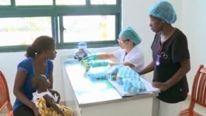 A Chinese pediatrician prescribes medication at the Sierra Leone-China Friendship Hospital | The Calabash Newspaper Mother with an infant sits beside a nurse who handles medical supplies at a clinic counter.