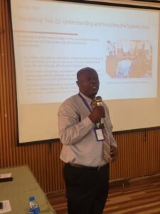 Executive Director of SLURC, Dr. Joseph Macarthy | The Calabash Newspaper Man in a light shirt and striped tie speaks into a microphone during a workshop, standing in front of a projected slide and wearing a lanyard badge.