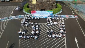 Aerial view of a large group of people in white shirts forming characters on a parking lot, with a blue banner and posters along a circular display