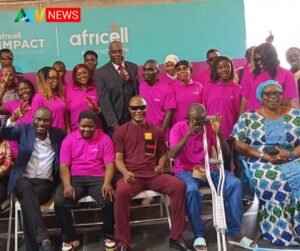 Group of people in pink shirts posing for a photo on a stage with a teal backdrop that reads Africa... News logo in the corner. central man with crutches sits front row.