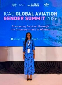 Woman in a blue floral dress stands in front of an ICAO Global Aviation Gender Summit 2026 backdrop with event slogan below the title.
