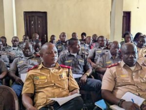 Group of uniformed officers seated in a hall, some wearing tan uniforms with red epaulettes, holding papers during a briefing or meeting.