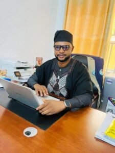 Man in a black traditional outfit and cap sits at a desk with an open laptop, wearing glasses and a smartwatch in an office.