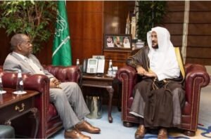 | The Calabash Newspaper Two men sit on red leather chairs in a formal office, facing each other as they talk; desk with a phone, framed photos, and a green flag nearby.