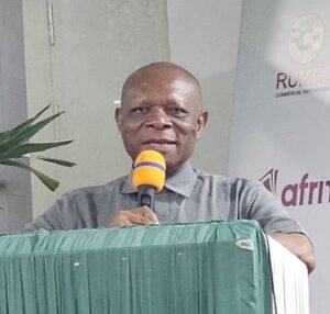 | The Calabash Newspaper Man in a gray shirt speaks into an orange microphone from a green podium at an event.