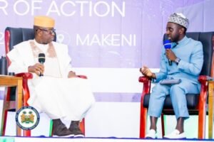 | The Calabash Newspaper Two men in traditional outfits sit on stage in red chairs, each holding a microphone, during a panel discussion against a purple banner backdrop (official seal visible on a podium).