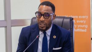 | The Calabash Newspaper Man in a navy suit and glasses speaks into a microphone at a conference, with an orange banner behind him.
