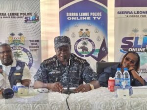 Sierra Leone Police officers sit at a press conference table with banners reading 'Sierra Leone Police Online TV' behind them; central officer speaks into a microphone.